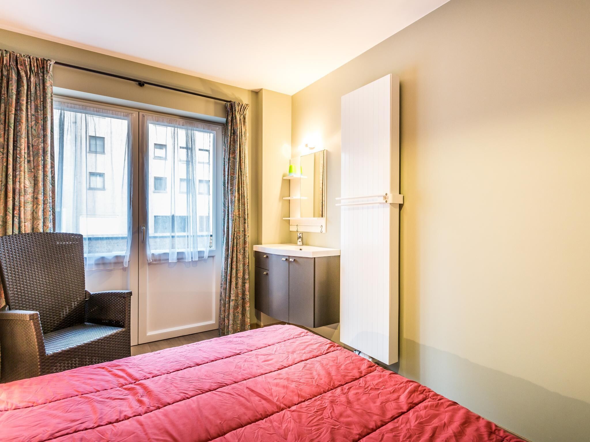 Habitación de hotel con cama, una silla, baño con lavamanos y espejo, y ventana con cortinas.