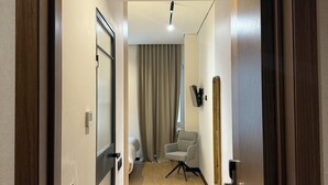 A hotel room with a wooden floor, a bed, a chair, a TV, and a window with curtains. - Grand Hotel Mosh Mosh (Almaty)