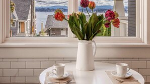 Quarto com vista pela janela para uma casa e um corpo de água. Há uma mesa branca com um vaso de flores, duas xícaras brancas e um jarro branco.