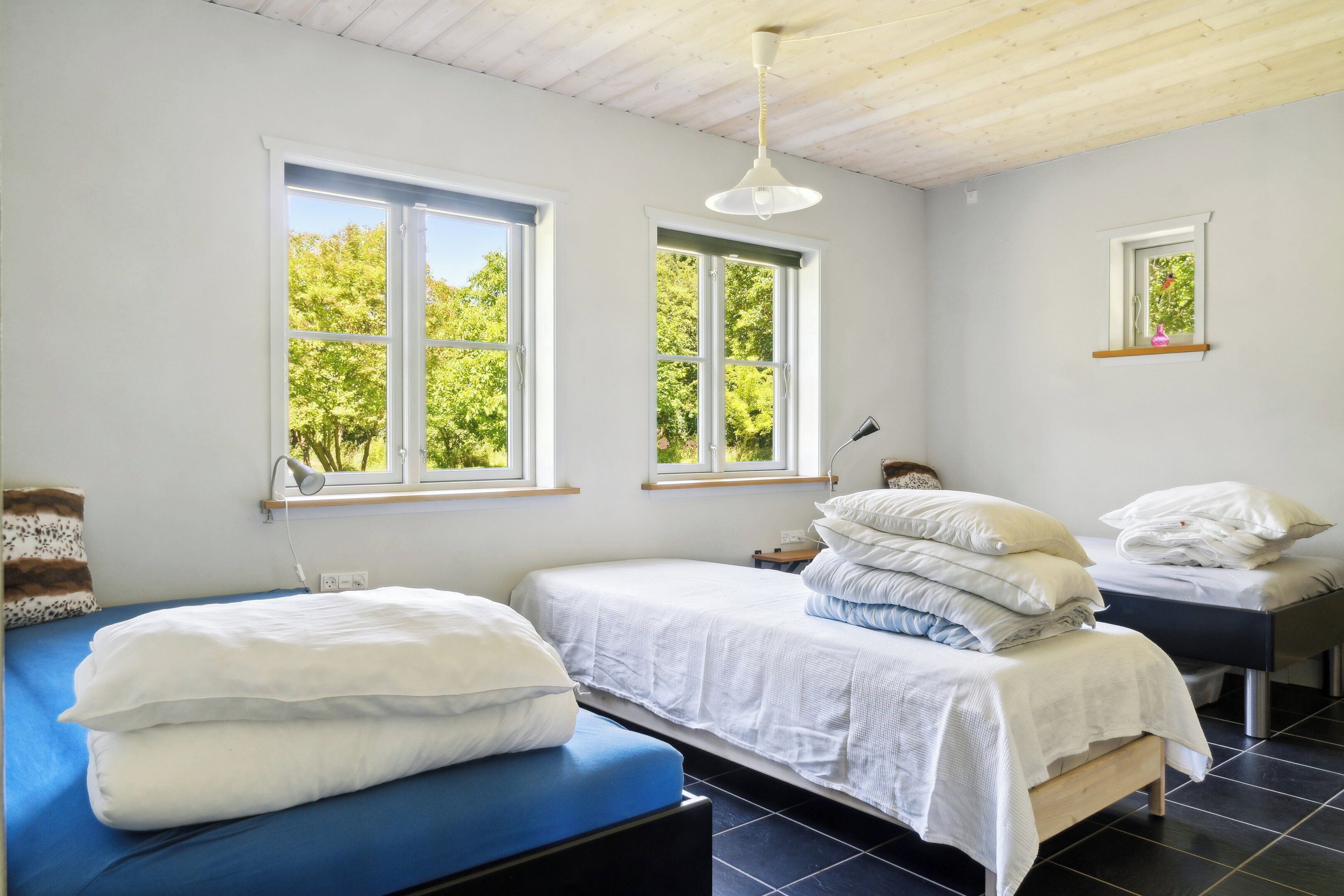 Quarto com duas camas, uma escrivaninha, uma cadeira e janela com vista para árvores.
