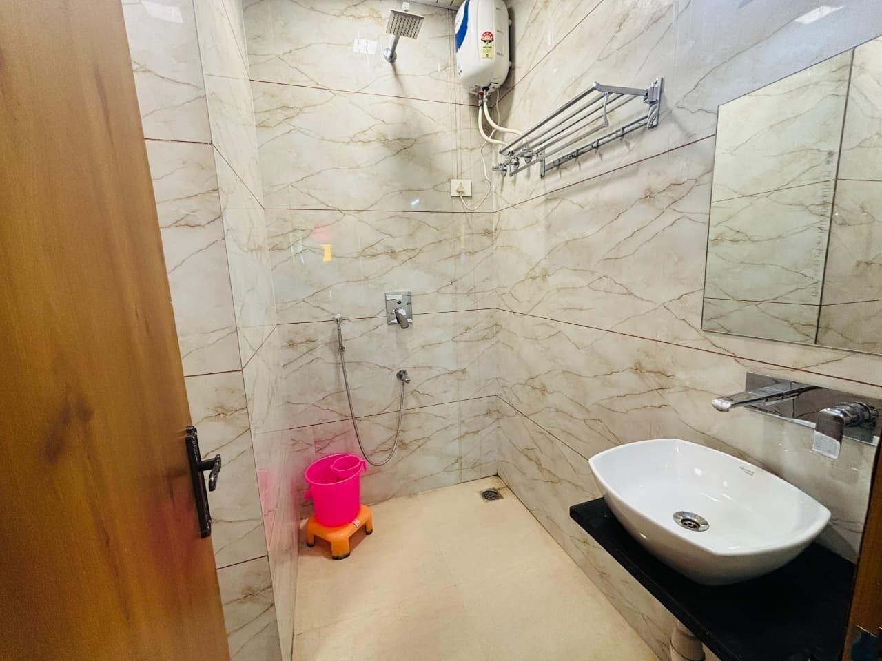 Un bagno con parete in marmo, lavandino bianco, doccetta e un secchio rosa su uno sgabello.