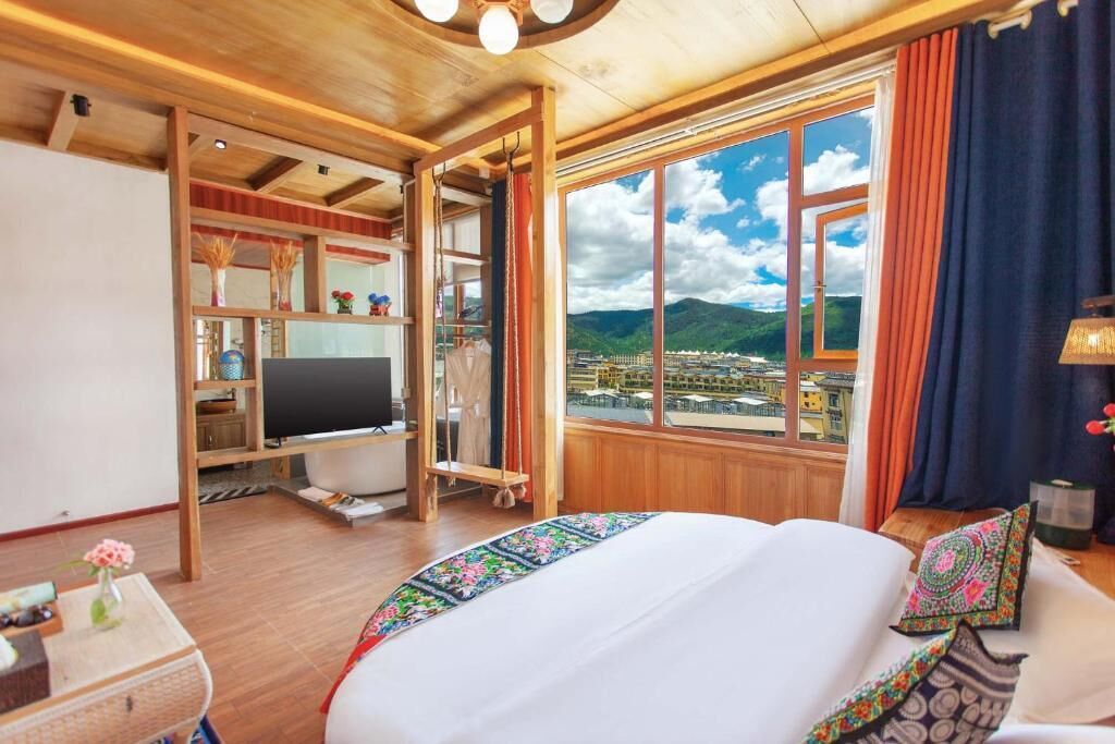 A hotel room with a large bed, a TV, a wooden desk, and a view of a cityscape through a large window.