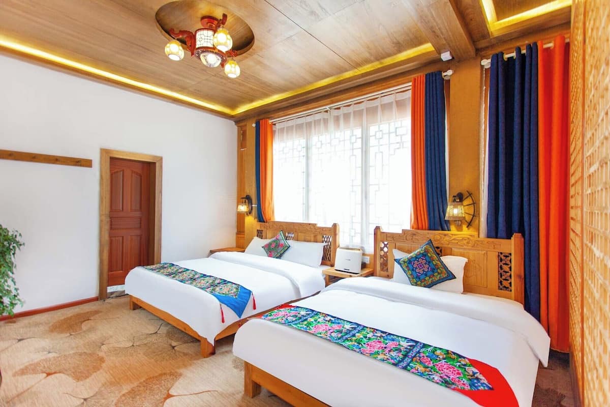 A hotel room with two beds, wooden headboards, a ceiling light fixture, a window with curtains, and a door.