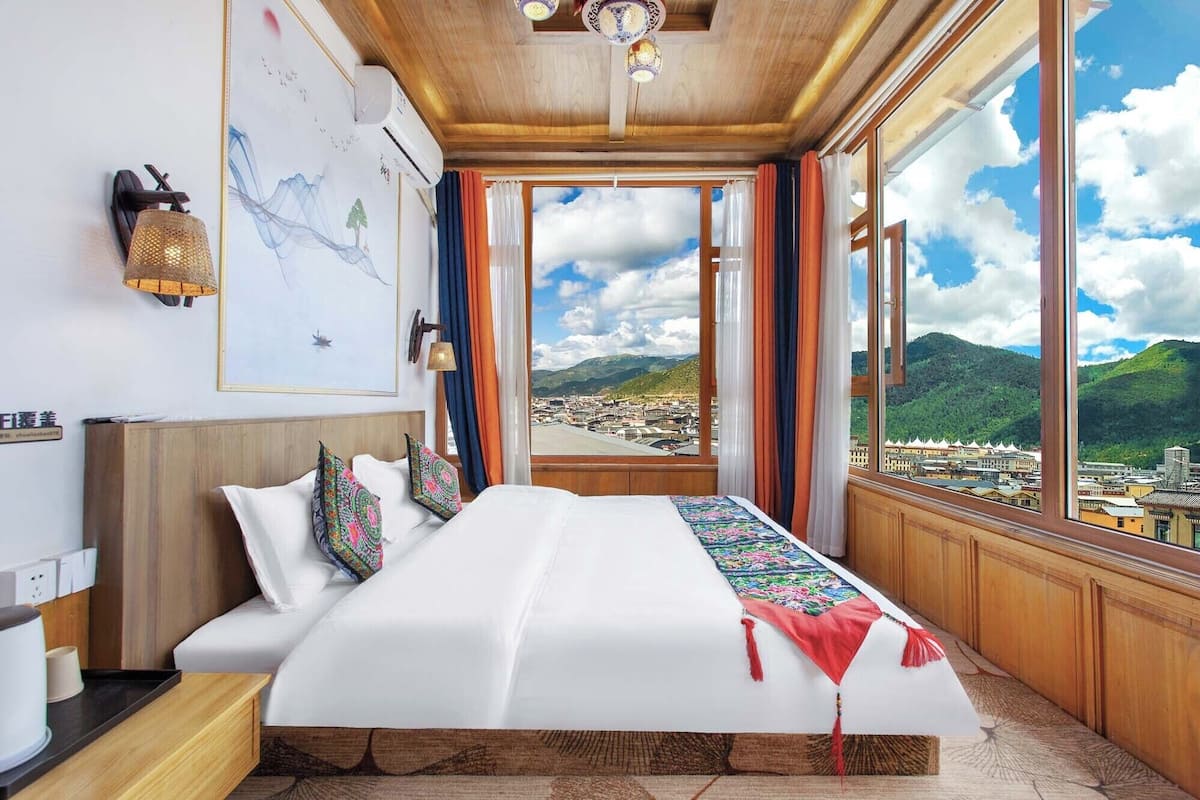 A bedroom with a large bed, a view of a town through a window, and a wall-mounted air conditioner.