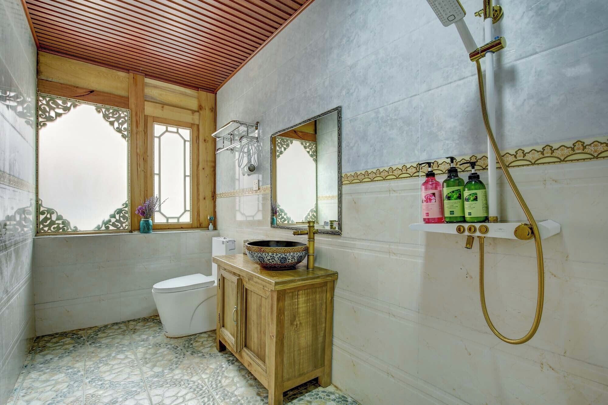 A bathroom with a wooden vanity, a white toilet, a large mirror, and a shower with a gold hose.