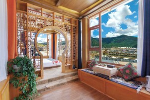 Una stanza con una grande finestra che offre vista su una città e montagne. Si vede un letto, una libreria in legno e un angolo salotto con cuscini.