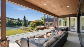 Un espace extérieur abrité, avec un canapé et une table en osier, une vue sur un lac et, au loin, un bâtiment moderne.