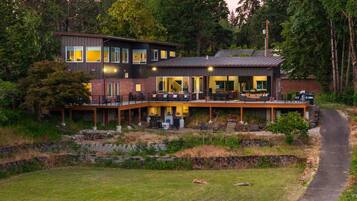 Une maison moderne à deux étages, dotée d’une terrasse et entourée d’arbres et d’une pelouse.