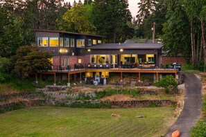 Une maison moderne à deux étages, dotée d’une terrasse et entourée d’arbres et d’une pelouse.
