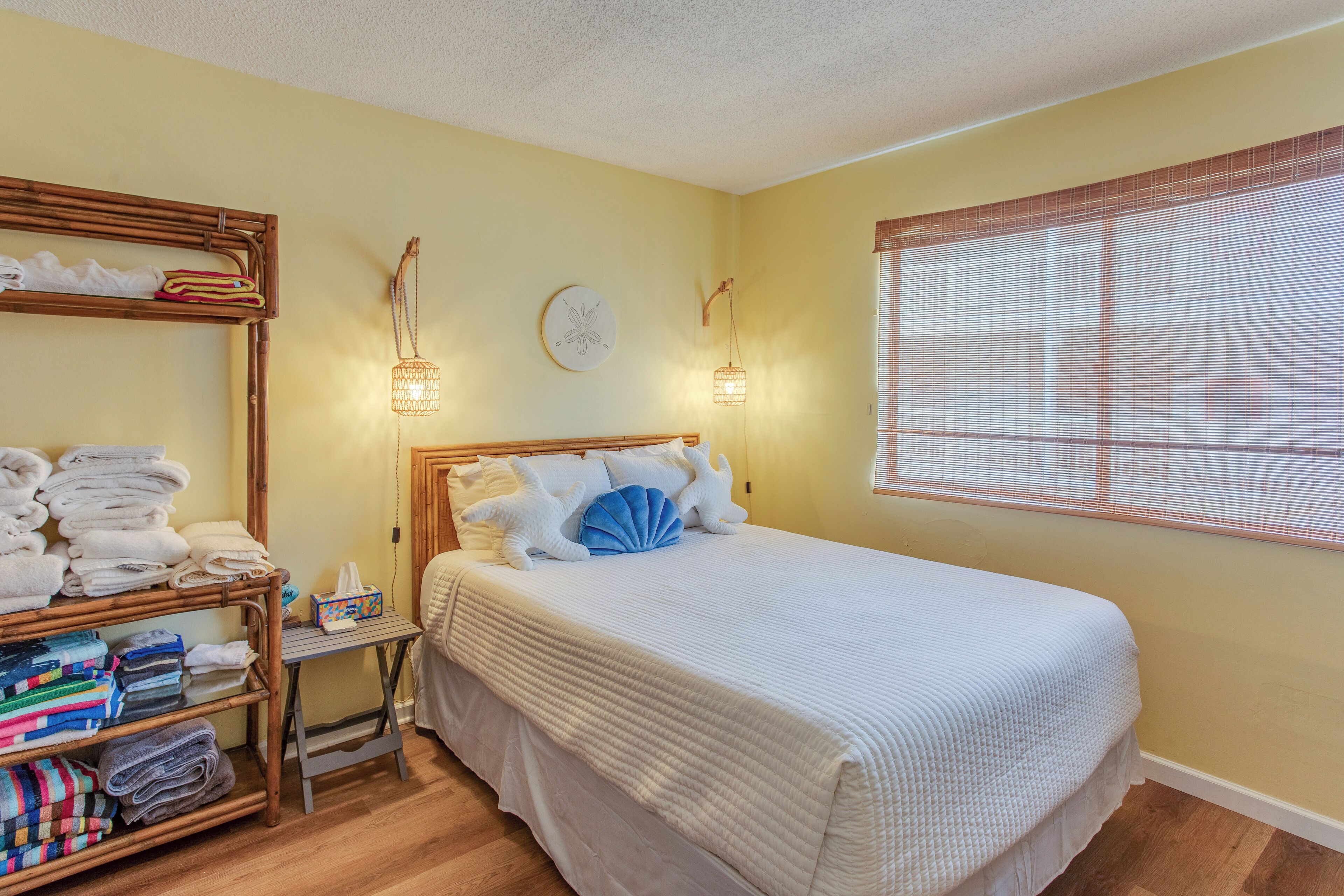 A neatly arranged bedroom with a bed, a shelf with folded towels, a bedside table, and a window with blinds.