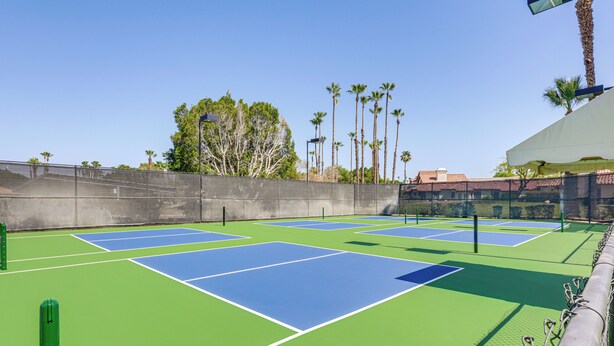 Un campo da tennis all'aperto con diverse superfici blu e verdi, circondato da una rete metallica e palme.