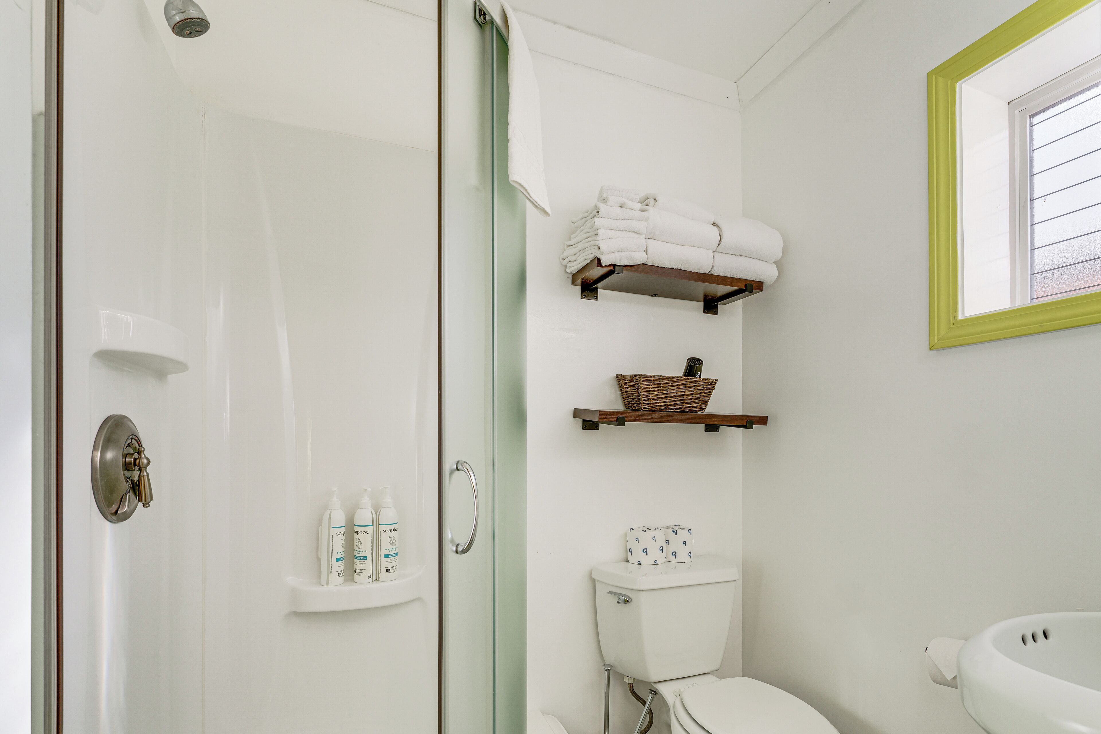 A bathroom with a glass shower door, a toilet, a sink, and shelves with toiletries.
