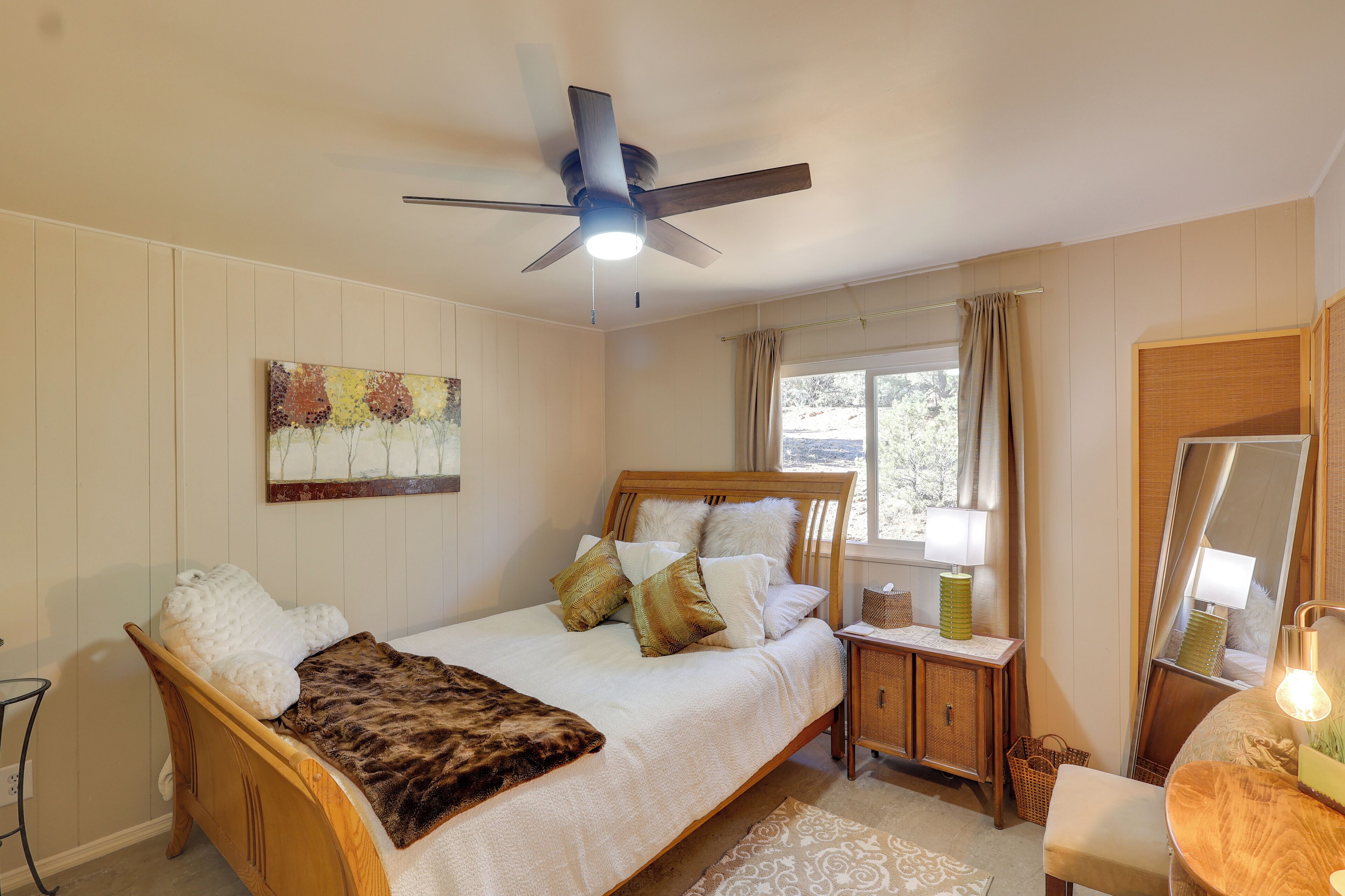 A bedroom with a wooden bed, a ceiling fan, a window with curtains, a bedside table with a lamp, and a mirror.