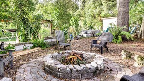 A stone fire pit surrounded by outdoor seating, including chairs and a bench, in a wooded area with trees and greenery. - Canal-Front Retreat w/ Hot Tub & Dock in Tavares (Tavares)