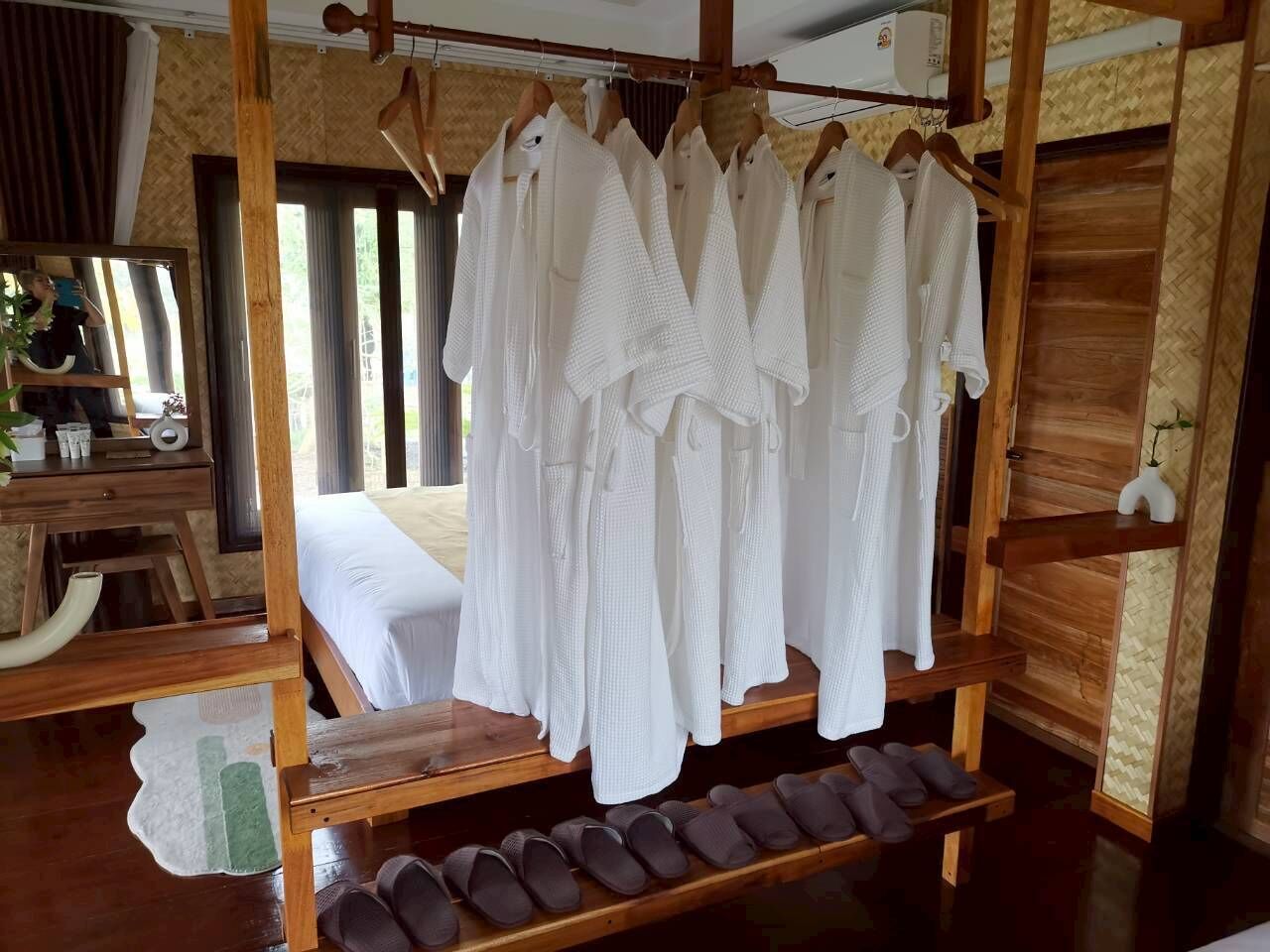 A wooden bed frame with white bedding, a wooden shelf with slippers, and a mirror reflecting a room with a window and curtains.
