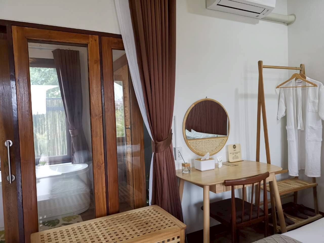 Una habitación con una puerta corredera de madera, un espejo redondo sobre una mesa de madera, un perchero blanco y una silla de mimbre.