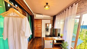 Een kamer met houten vloer, een houten garderobe, een flatscreen televisie, een kleine koelkast en een raam met uitzicht op groen.