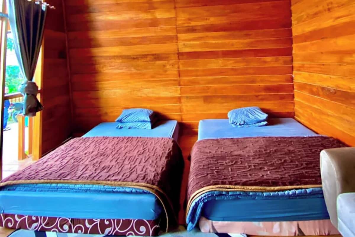 Quarto de cabana de madeira com duas camas, um candeeiro pendente e vista para o exterior através de uma janela.