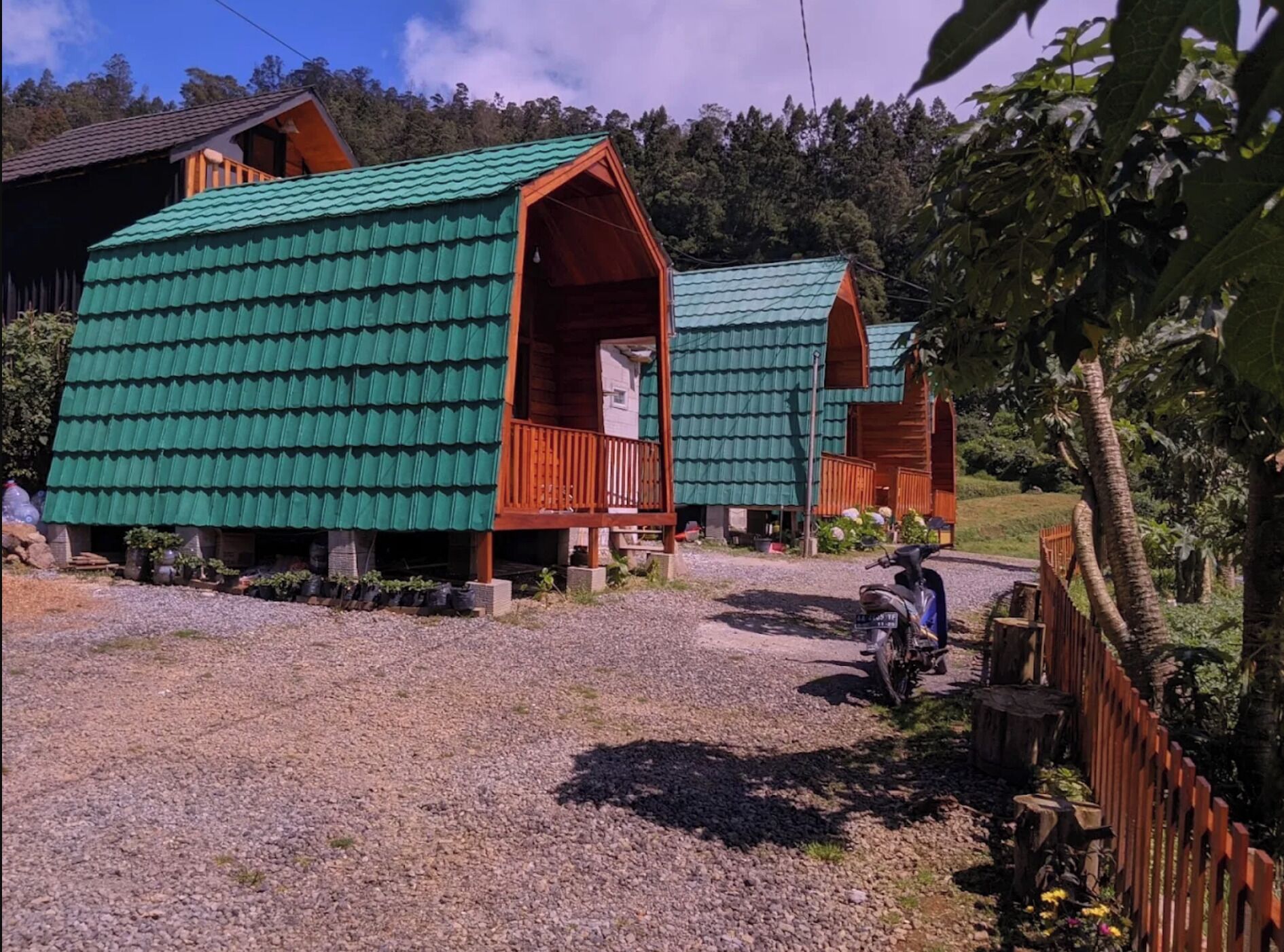 Filas de cabañas pequeñas con techos verdes y balcones de madera, ubicadas en una zona rural con senderos de grava y rodeadas de árboles.