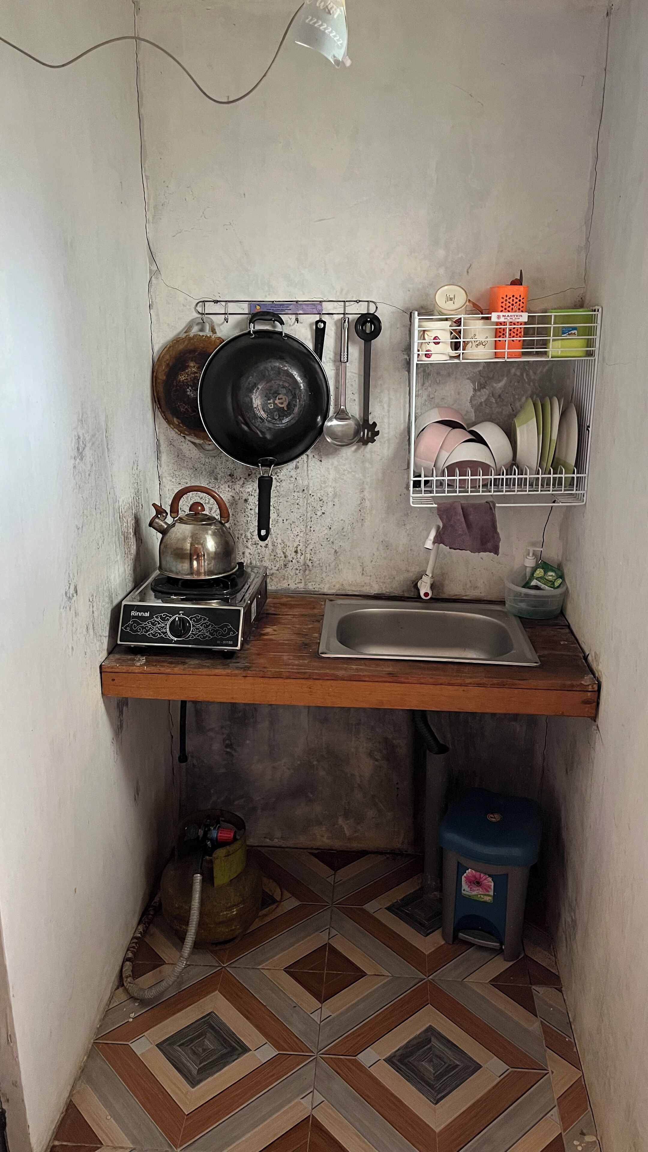 Une petite cuisine équipée d’une cuisinière à gaz, d’une bouilloire, d’une poêle, d’une louche et d’une étagère contenant de la vaisselle et un récipient.