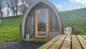 A rustic wooden structure with a door, a small garden area with a wooden bench, and a pair of glasses on the bench. - Birds pod is the perfect place for up to two guests to reconnect with nature. (Dalton-in-Furness)