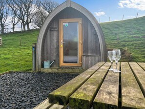 Une structure rustique en bois, dotée d’une porte, d’un petit espace jardin avec un banc en bois, et d’une paire de lunettes posée sur le banc.