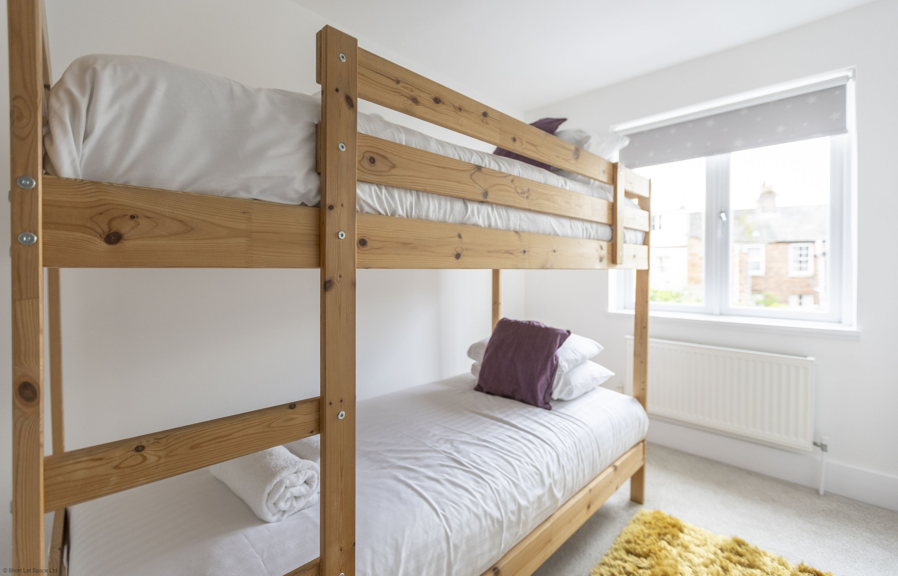 A bunk bed with white bedding and a purple pillow, a window with blinds, and a radiator.