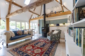 A spacious living area with a high ceiling, exposed wooden beams, a large rug, a sofa, a patterned armchair, and a bookshelf. - The Cart Shed | Tackley Holiday Let | 2 Beds & Sleeps 4 (Tackley)
