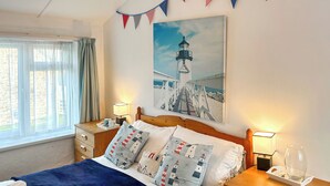 Una cama bien hecha con un edredón azul, dos mesitas de noche con lámparas, un cuadro de un faro y una ventana con cortinas.