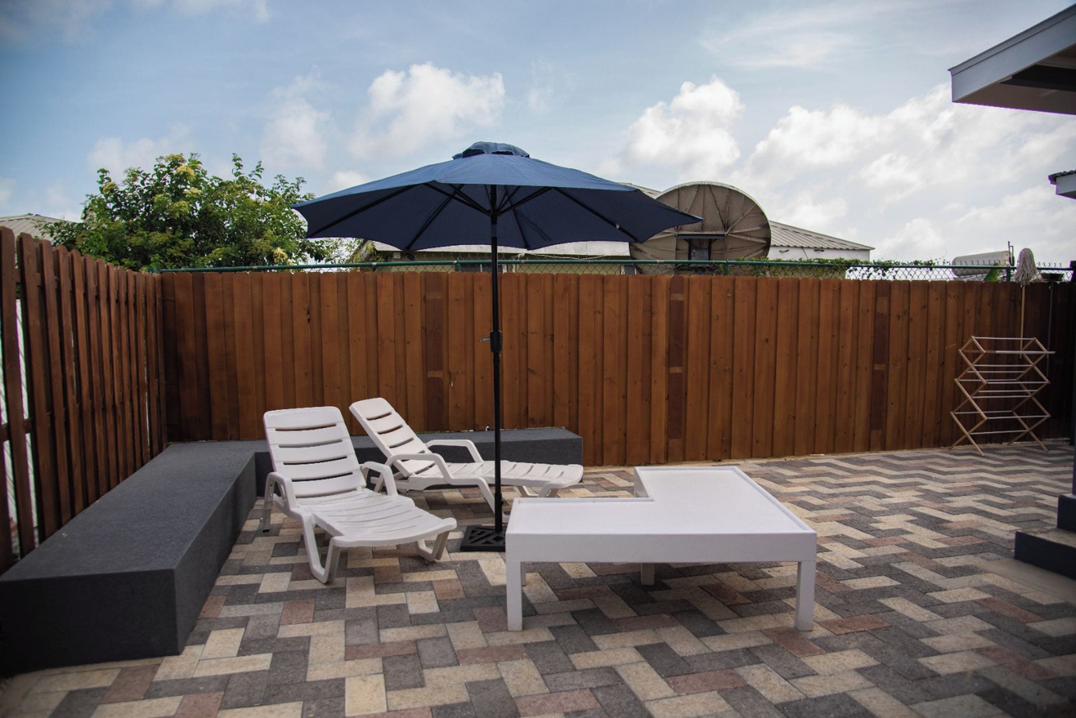 Une terrasse extérieure avec une clôture en bois, un sol carrelé à motifs et un espace détente blanc comprenant une table et des chaises.