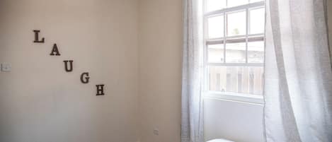 Ein Schlafzimmer mit einem Bett, einem Fenster mit Vorhängen und den Buchstaben ‚LAUGH‘ an der Wand.