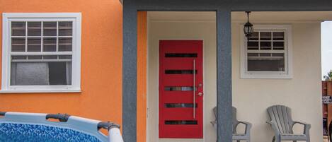 A swimming pool, a red door, and two Adirondack chairs outside a building.