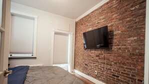 A bedroom with a brick wall, a flat-screen TV mounted above the bed, and a window with blinds. - Prime East Village Two Bedroom (New York)