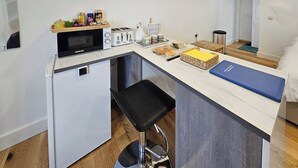 Et lille tekøkken med mikrobølgeovn, brødrister og et lille køleskab. En barstol står under en disk, og der er forskellige madvarer samt en blå bakke på disken.