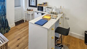 Et kompakt køkken med en mikrobølgeovn, toaster og kaffemaskine på bordet. Der er et lille bord med en kurv og en blå mappe.