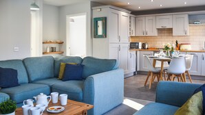 Un salon moderne avec un canapé bleu, une table basse en bois sur laquelle sont posées des tasses et une plante, et une cuisine en arrière-plan avec des meubles blancs et un four à micro-ondes.