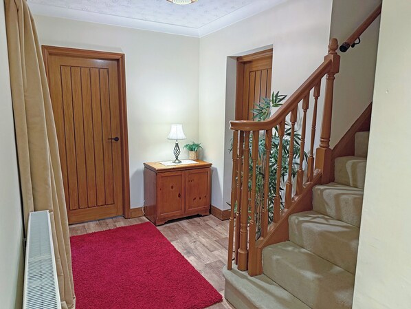 Un pasillo con puertas de madera, un tapete rojo, una lámpara, un mueble pequeño y una escalera de madera.