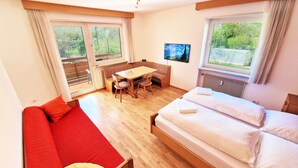 Une pièce comprenant un lit, un canapé rouge, une table à manger en bois avec des chaises, et un téléviseur fixé au mur.