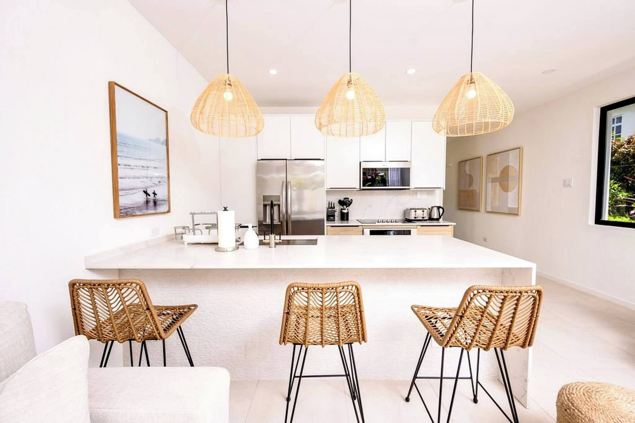 A modern kitchen with a large island, wicker chairs, and pendant lighting.