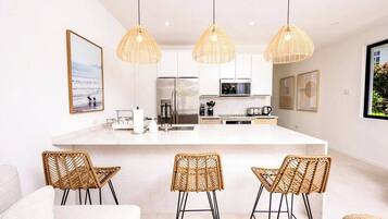 A modern kitchen with a large island, wicker chairs, and pendant lighting.