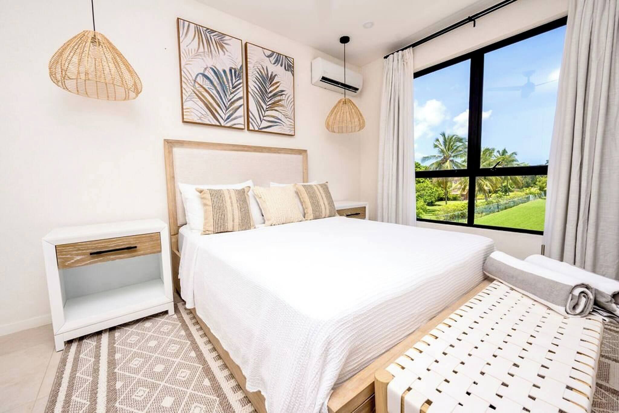 A bedroom with a large bed, a nightstand, a window with a view of greenery, and two pendant lights.