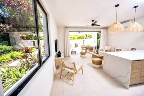 A modern kitchen with a large island, a dining area with a table and chairs, and a view of a garden through a large window. - Azzurro Townhouses - by Zenbreak (Speightstown)