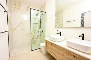 A modern bathroom with a glass-enclosed shower, a white toilet, and a wooden vanity with two sinks. - Azzurro Townhouses - by Zenbreak (Speightstown)