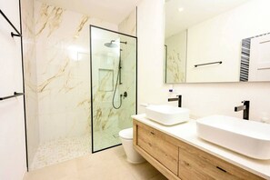 Un baño moderno con ducha de vidrio, inodoro blanco y tocador de madera con dos lavamanos.