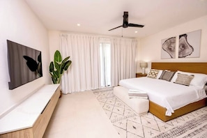 A modern bedroom with a large bed, a ceiling fan, a TV mounted on the wall, and a large window with curtains. - Azzurro Townhouses - by Zenbreak (Speightstown)