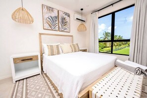 A bedroom with a large bed, a nightstand, a window with a view of greenery, and two wall-mounted lights. - Azzurro Townhouses - by Zenbreak (Speightstown)