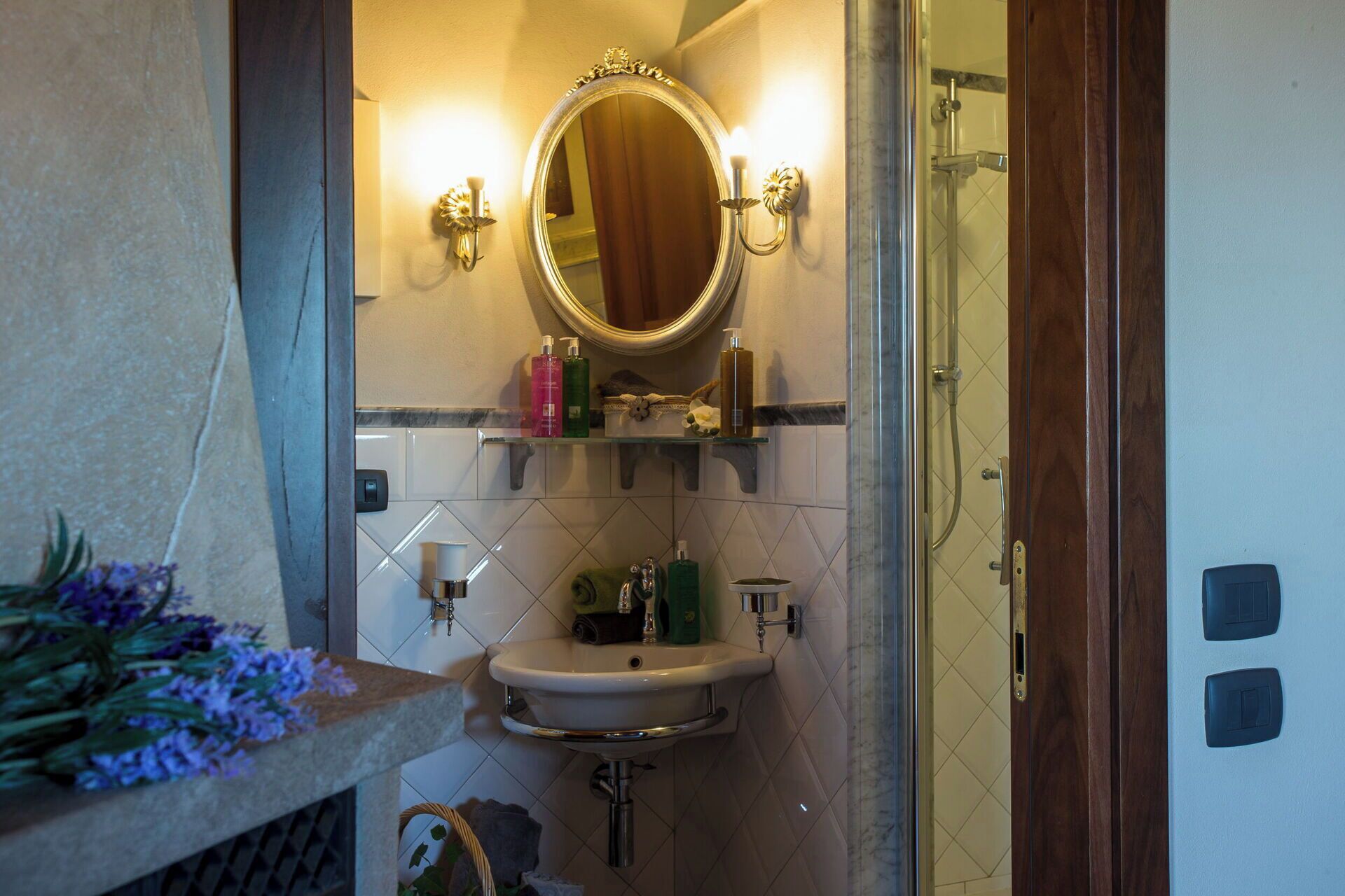A bathroom with a sink, mirror, and shower. There are toiletries on the wall and a vase with flowers on the counter.