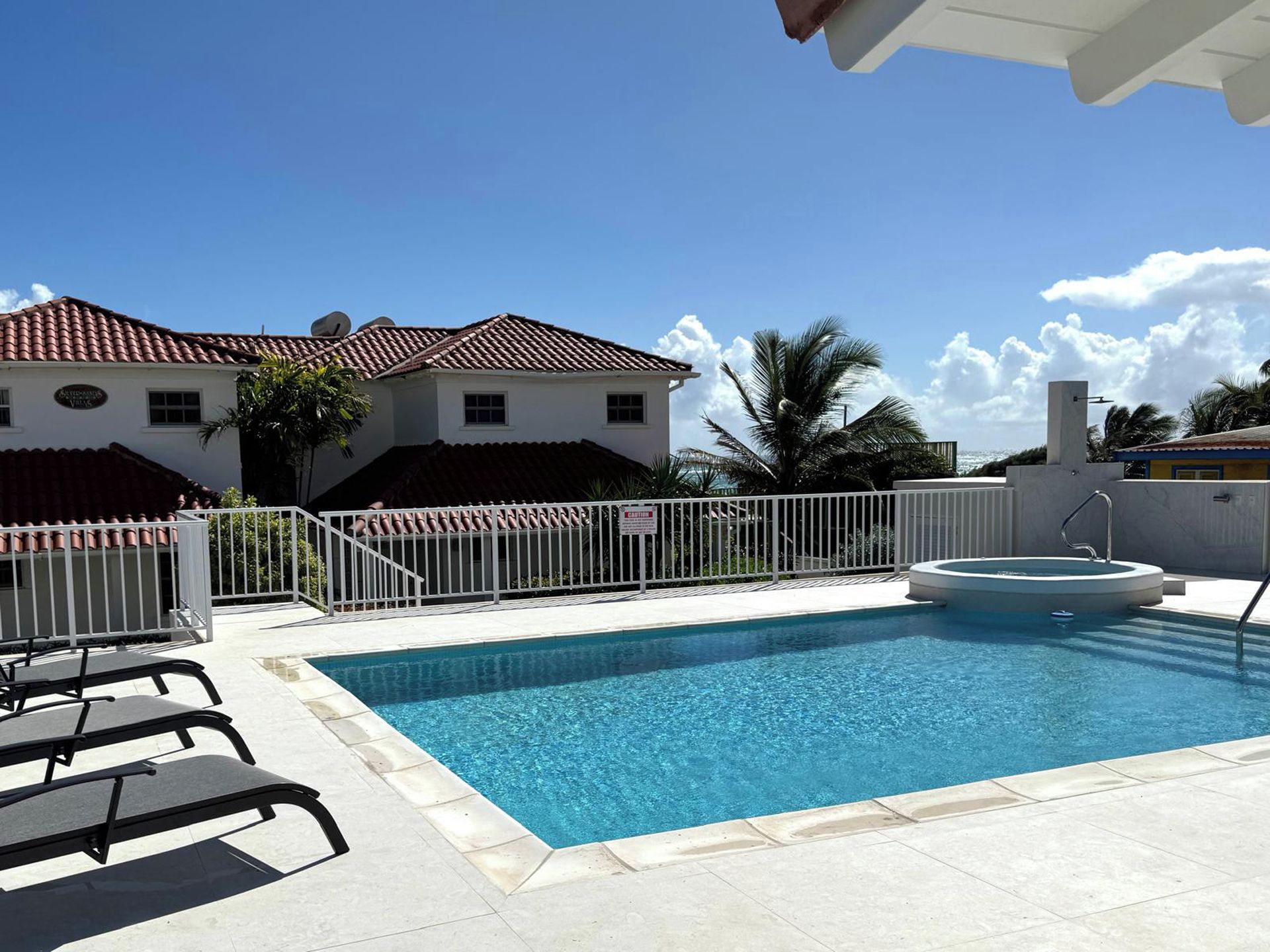 A swimming pool with a white fence, lounge chairs, and a building with a red-tiled roof.