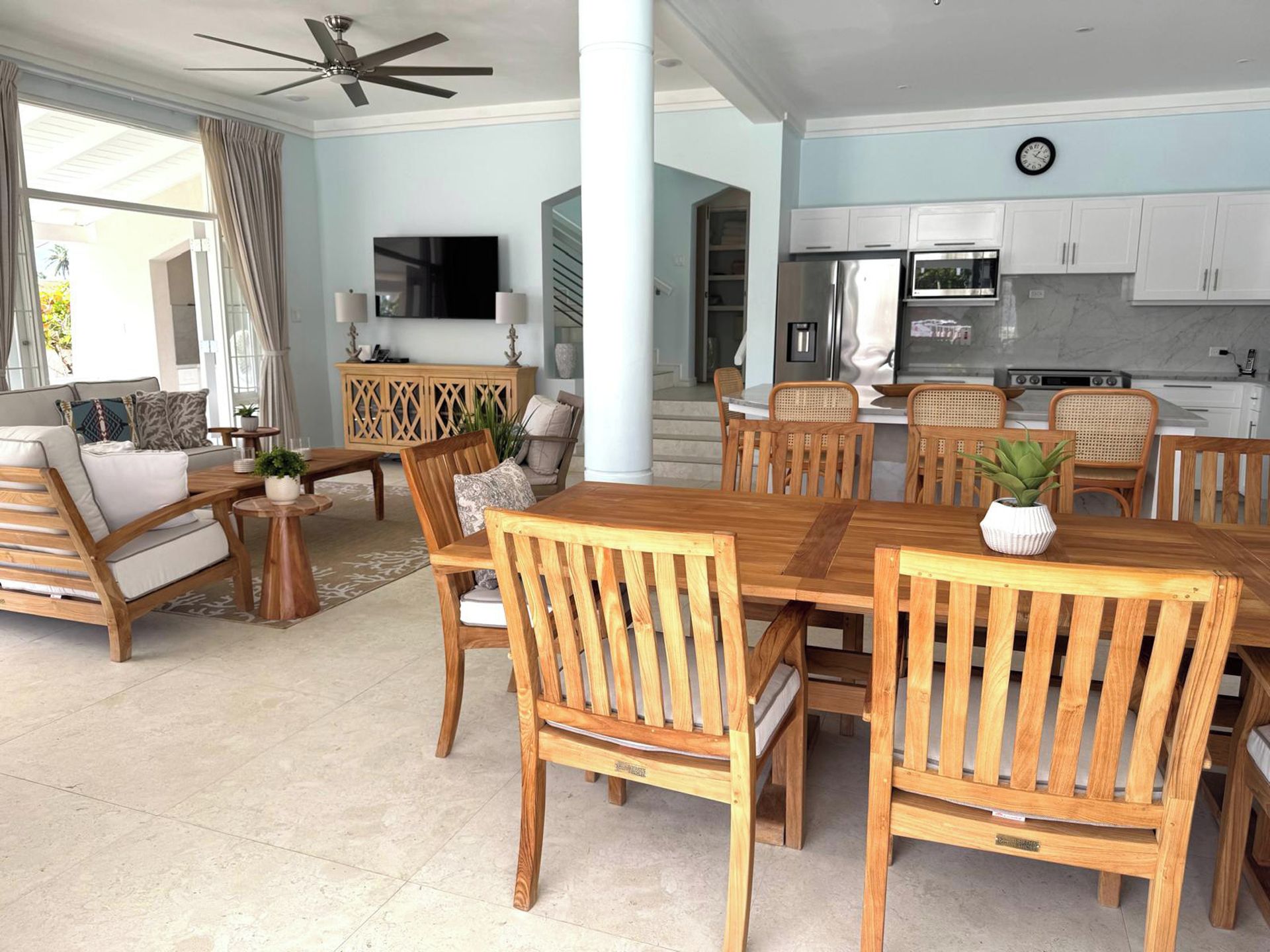 A spacious living area with a wooden dining table, chairs, and a sofa. A TV is mounted on the wall, and there is a kitchen area with cabinets and appliances.