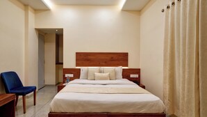 Camera d'albergo con un poggiatesta in legno, un letto grande con biancheria bianca, una sedia blu, un comodino con una lampada e una tenda alla finestra.
